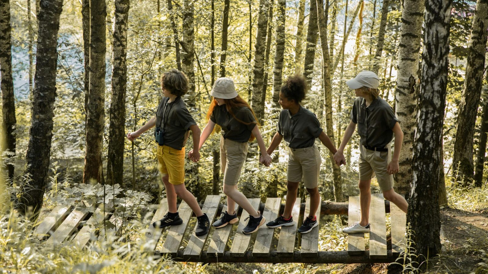 A group of teenagers walk hand in hand across a wooden bridge in a sunny forest.